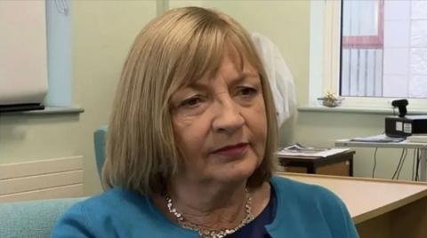 Lesley Walker is wearing a blue jacket and black top. She has shoulder length blonde hair with a fringe and is sitting in an office.