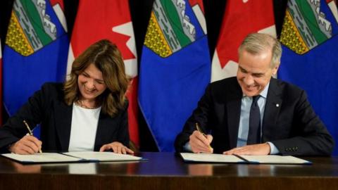 Canada's Prime Minister Mark Carney and Alberta's Premier Danielle Smith sign a Memorandum of Understanding (MOU), before the Prime Minister's energy-related announcement in Calgary, Alberta, Canada November 27, 2025. They are seated, and pictured each signing a document. Behind them is a row of Canada and Alberta flags. 