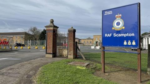 A blue RAF Scampton sign stands at the entrance to the site with an RAF logo and the badge of RAF Scampton. There are two brick gate posts at the entrance and buildings in the background