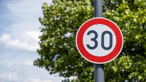 Generic image of a circular 30 mph sign with greenery and blue skies in the background.