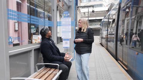A black man sat a tram stop in a black coat and hat. He is talking to a female BBC reporter with blonde hair. She is wearing a black gillet and faded blue jeans. A blue Midlands Metro tram is passing by