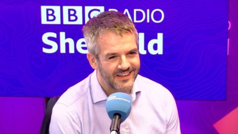 A man has short cropped grey hair and stubble and is wearing a white open necked shirt. He is sitting in a radio studio with a blue microphone in front and the words BBC Radio Sheffield on a purple wall behind him