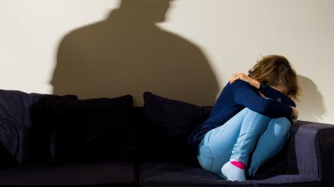 A woman in a blue jumper and jeans cowers, with her face in her hands, in the corner of a navy sofa while a shadow of a man towers above her.