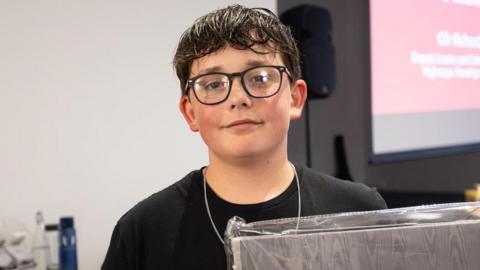 Matthew smiling at the camera, while standing in a room with a screen and a speaker behind him. He has short dark hair and glasses and is wearing a black T-shirt.