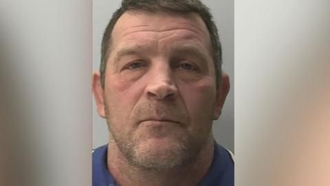 A mugshot of Paul Quinn, who is wearing a blue top, has light stubble, and is staring vacantly forward