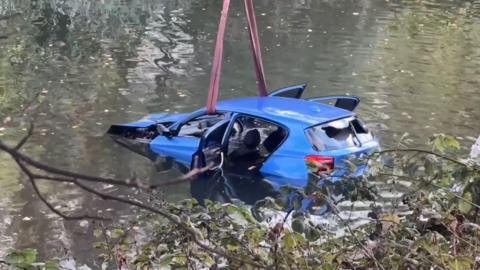 A blue car, with three open doors and a smashed rear window is submerged in river water. Red straps are looped through the driver and passenger doors to pull it out of the water.