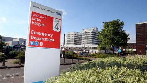 The Lister Hospital main buildings seen from an entrance with a white and red sign saying "Welcome to Lister Hospital Gate 4 Emergency Department". In the background are the grey multi-storey hospital buildings  and in the foreground is the drop-off point with a bus and some foliage and trees.