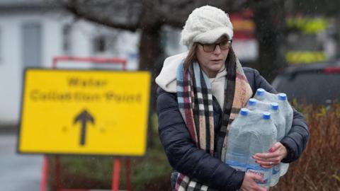 Woman carrying bottles of water in rain. In background is a sign for a water collection point