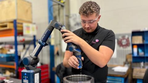 A young person wearing safety glasses uses a portable measuring arm to inspect a large metal component in a workshop. Industrial shelving, tools and equipment are visible in the background.