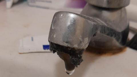 Close-up of a corroded bathroom tap with a droplet of water hanging from the spout, indicating a leak. The metal surface shows signs of rust and mineral buildup. A toothpaste tube and packaging are partially visible in the background.