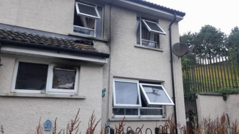 Shows a house with scorch damage and windows open with a satellite dish to the right and, to the right of that, fencing, a grass verge and trees.