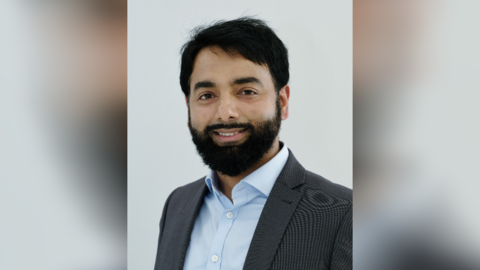 Ejaz Ahmed is stood in front of a grey background and is wearing a light blue buttoned shirt, with a grey jacket. He has short black hair and a black beard