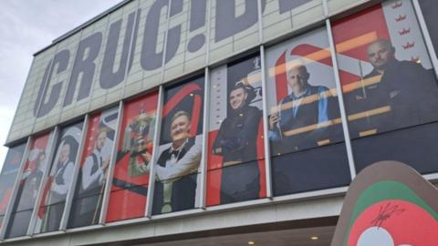 The exterior of the Crucible Theatre has white tiles and large, long windows. In each of the windows are large posters of different snooker players with black, white and red branding
