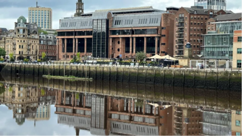 A large, pink building with black windows sitting behind a river.