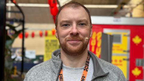 James is smiling at the camera. He is wearing a grey t-shirt and hoodie and has an orange lanyard around his neck. 