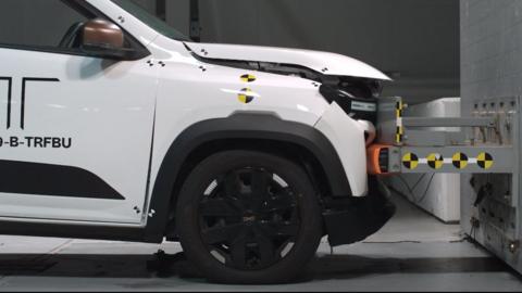 A white electric car crashing into a wall at a low speed in a crash test lab. The front of the car has bent in a bit and the bonnet has lifted up, but otherwise the damage looks minimal.