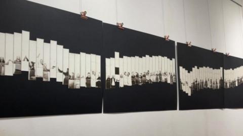 Four black panels hanging on the wall, each partly covered with about 20 thinly cut photographs of people, taken in black and white.