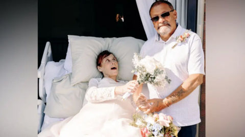 Karen and Stephen Glossop dressed in white holding flowers in a hospice bed