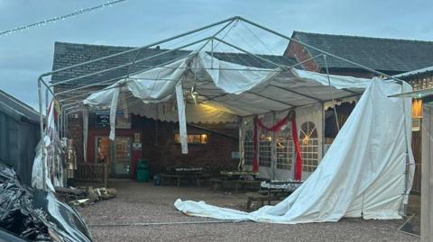 A damaged marquee with no roof on the top. The fabric is hanging off the poles, with Christmas decorations still in place.