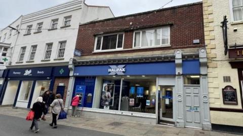 Buildings on Baxtergate in Whitby. The centre building has a blue Halifax bank sign above a large glass front window. There is a shop to the left hand side and a pub sign on the building on the right. There are some people walking past the front of the buildings.