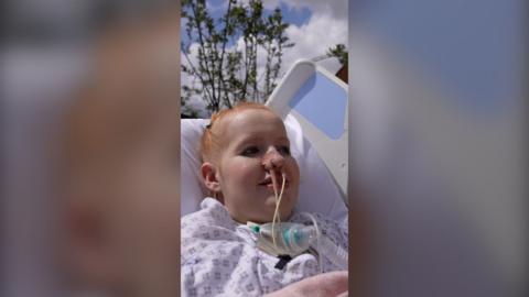 A woman with ginger hair lies in a hospital intensive care bed. There are tree branches and blue sky behind her.