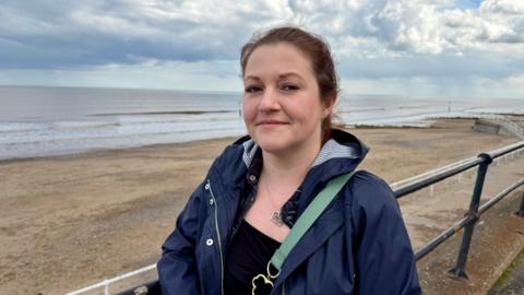 A woman with brown hair, which is tied back. She is wearing a black top and a navy coat. She is standing close to the railings on Hornsea seafront with the sea in the background. She is smiling at the camera.