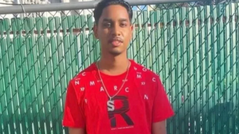 A young man in a red t-shirt and a silver chain with the letter s on, looking at the camera