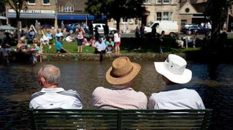 A photo of three men sitting on a bench with their backs to the camera. They are sitting in front of a stream with people in the background. Two of the men are wearing sunhats.