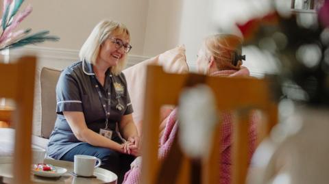 Stock image of a female nurse chatting to another woman. The nurse has shoulder-length blonde hair and a fringe, and is wearing glasses and a navy uniform. She is sitting on a sofa as she talks to the fair-haired woman, who is wearing a pink dressing gown. They are in a living room and biscuits and a cup of tea are placed on a small table next to the pair.