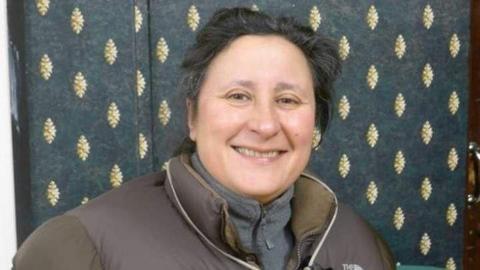 A woman with short black hair smiles at the camera. A gold-coloured earring hangs on her left ear and behind her is a curtain with a gold leaf-like design on a dark background. She wears a puffy brown jacket over a grey fleece top