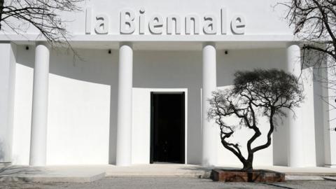 A white building bearing the words "la Biennale" with a black tree in front of it.