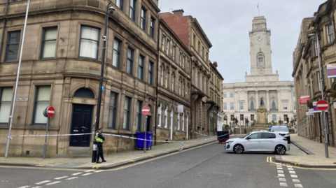 A street in Barnsley, with a police officer standing guard on the corner. A police cordon extends around a pavement on the left. A grand civic building is visible in the distance.