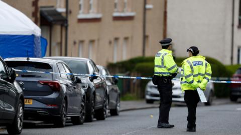 A male and a female police officer standing in front of a line of blue and white police tape blocking off a street. Parked cars, homes and a blue and white forensic tent can be seen on the left of the image.