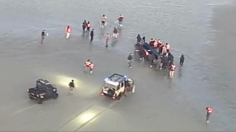 Two cars in the shallow water with people surrounding what appears to be a dingy boat with people inside the vessel