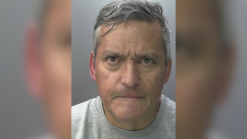 A man with short grey hair and a stern expression wearing a grey t-shirt in a police custody picture.