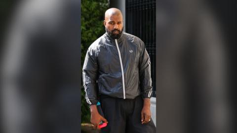 Kanye, or Ye, West has a shaved head and a black bear. He is a wear black trousers, and a black windbreaker with a white zip.