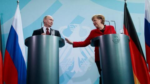 Putin and Merkel stand at cylindrical podiums smiling at each other, at a news conference in Berlin in 2012. Merkel is holding out her right hand towards Putin.