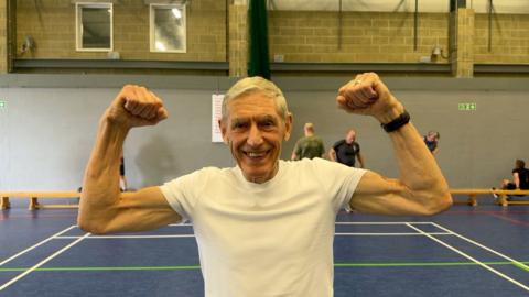 A man is stood in a gymnasium flexing his muscles. He is wearing a white t-shirt and there are people training behind him. 