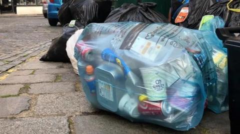 A blue bag, on a pavement, filled with rubbish. There are black bags around them. 