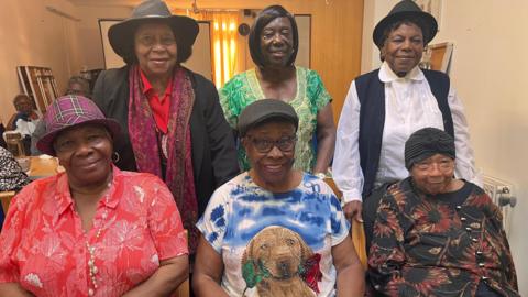 Group of women stood in a group wearing colourful clothing, all in their 70s and 80s and 90s.