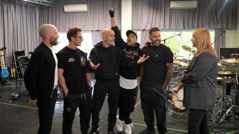 The five members of Five, all dressed in casual black clothing, stand in a rehearsal studio talking to a BBC journalist