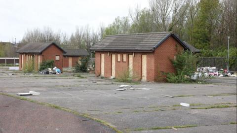 Two abandoned brick buildings, with bordered up windows. Rubbish sits behind them. 