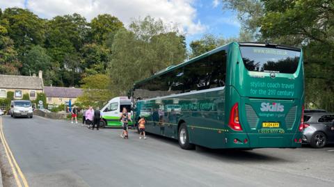A big green coach in a parking area in Bibury. It is a narrow area with small cottages and other cars surrounding it.