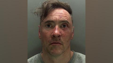 A police custody photo of Paul Doyle wearing a grey jumper and looking stunned. His hair, a long brown fringe, is dishevelled and to the side. His face has red marks on it