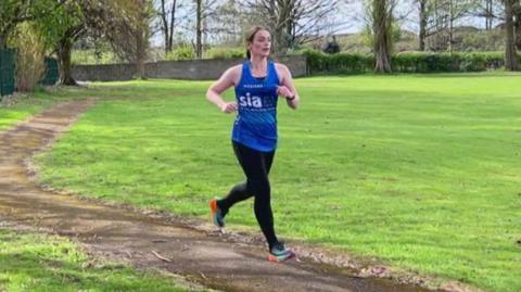 Emma Watt wearing a blue top running in a park with trees behind her.