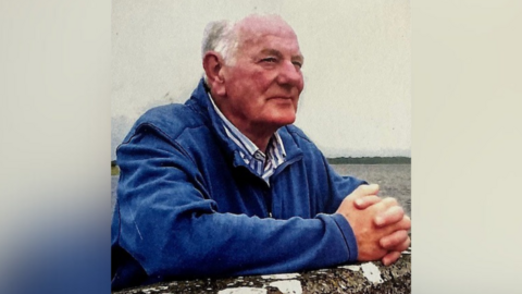 A picture of a man with white hair looking to the side, he is wearing a blue jacket and white and blue striped shirt, his arms are leaning on the wall.