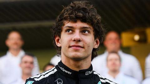 Kimi Antonelli as a racing driver looking up, with a black racing top, with people behind him, blurred. He has short dark curly hair. 