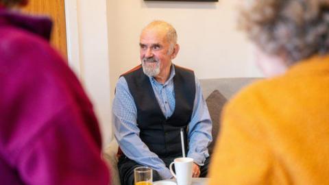 An older man with grey hair and facial hair, wearing a blue shirt and black waistcoat. He is looking to the side and smiling. In the foreground there are two people blurred - one in a pink top and another in a yellow top with curly grey hair