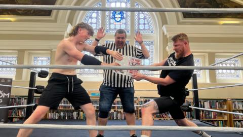 A boy in black shorts and no shirt stands on the left, a man wearing black shorts and a black T-shirt is on the right. They are both holding onto a book, fighting over it. Behind them stands a ref. All three are standing in a boxing ring. Behind them is a large, arched stained glass window above bookshelves.