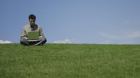 A young man is sitting cross-legged on the brow of a grassy hill typing on a laptop. He is wearing trousers, a shirt and a tie and his head is bowed as he types. It is a clear blue sky with a couple of wispy clouds.